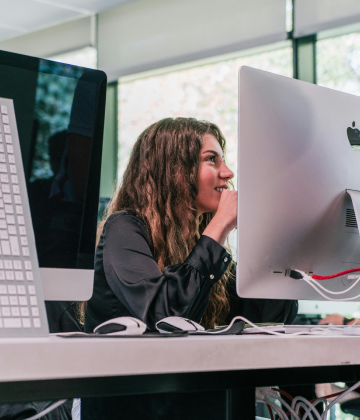 Experiencia transformadora - Aprende Haciendo En un aula de UNIE, vemos una alumna sonriente que atiende la clase y trabaja con un mac de pantalla grande.